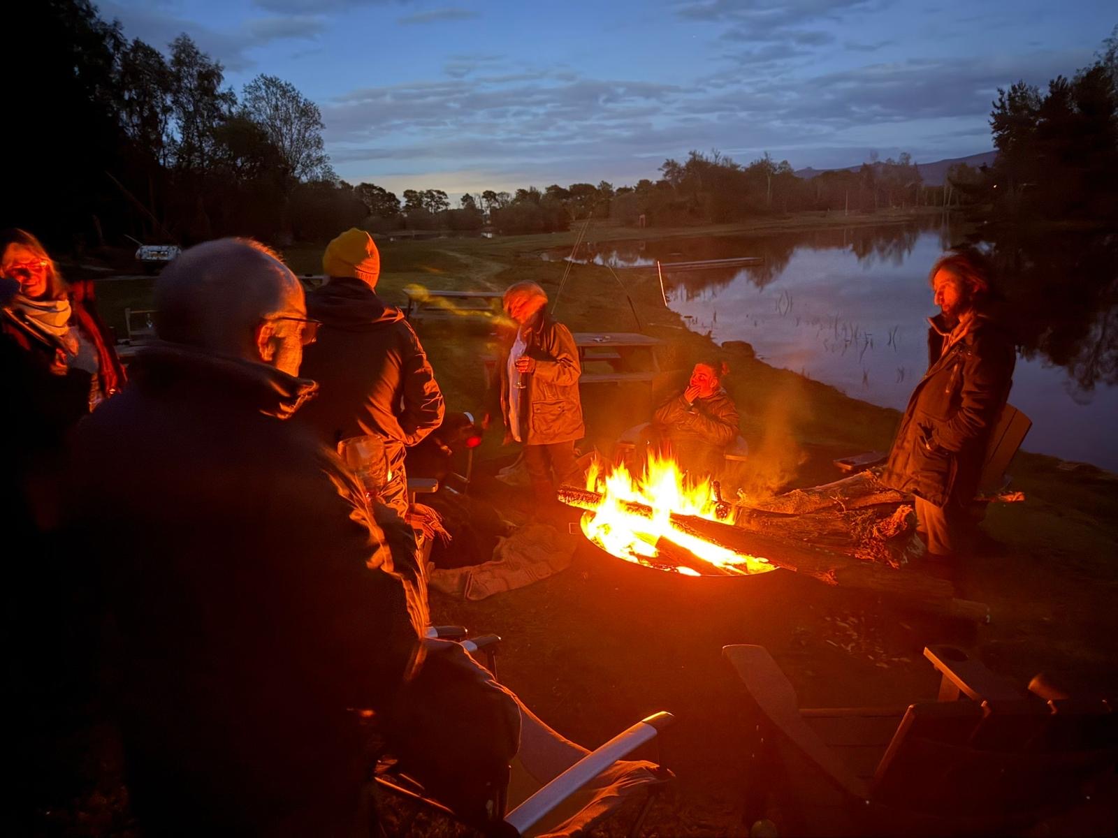 Group of visitors gathered around a campfire on the lochside at dusk
