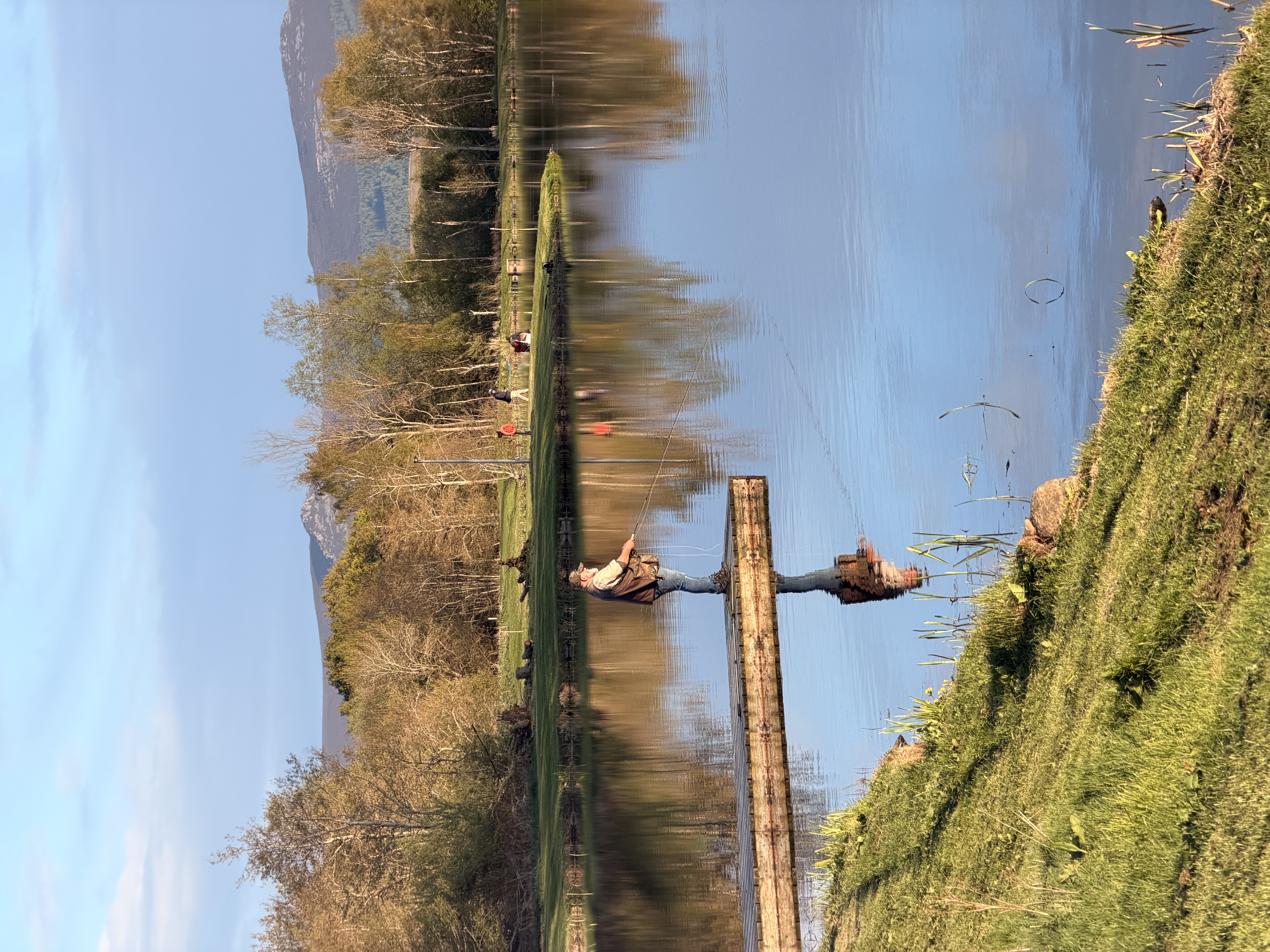 Angler casting a fly line from the jetty on a bright spring morning with Bennachie reflected in the loch